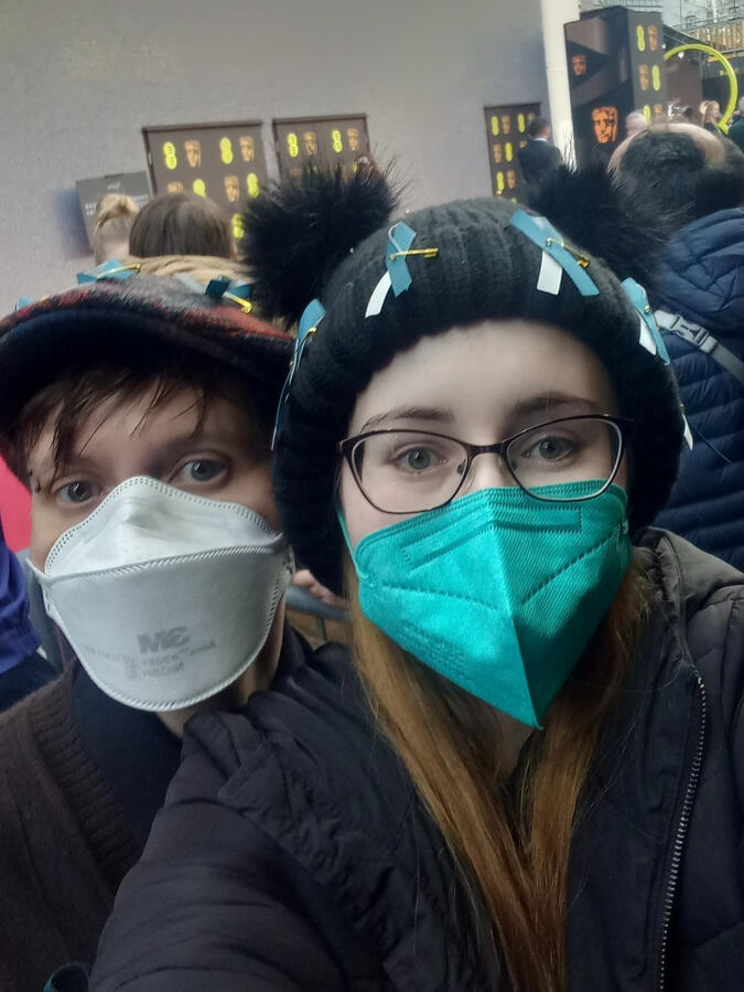 Two Protect the Heart of the arts campaign members, one wearing a white respirator, the other wearing a teal respirator to match Long Covid awareness ribbons on their beanie