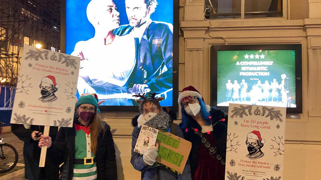 West End Festive Mask Handout -- four people in holiday costumes wearing masks and holding up signs, offering free masks outside the theater