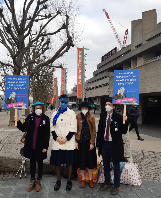 Four people wearing masks and holding up signs advocating for accessibility improvements outside the National Theater in London
